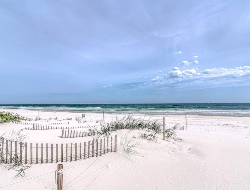 A sandy beach landscape is featured, with soft white sand and gentle dunes lined with small wooden fences. Grassy vegetation is seen among the dunes, while the calm turquoise waters of the ocean extend into the horizon under a sky dotted with wispy clouds.