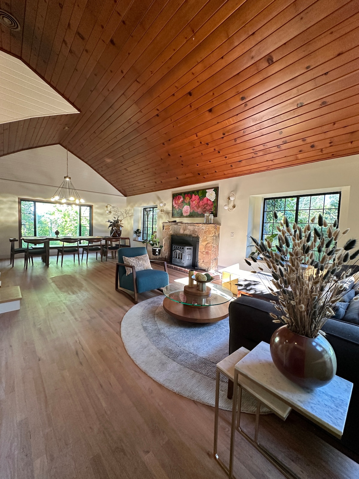 A spacious living area features a high, wooden-beamed ceiling and large windows framing views of the surrounding nature. A round coffee table is positioned on a soft area rug, complemented by contemporary seating options. A fireplace adds warmth and charm to the room.