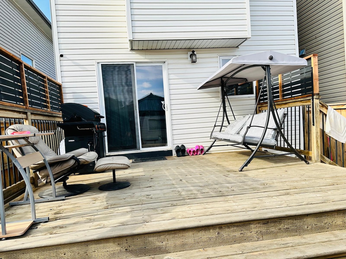 A deck area features a comfortable swing bed under a canopy, providing a relaxing space. Nearby, a grill is positioned, and a few seating options are arranged, including a lounge chair and a small table. The deck offers access to the home's entrance.