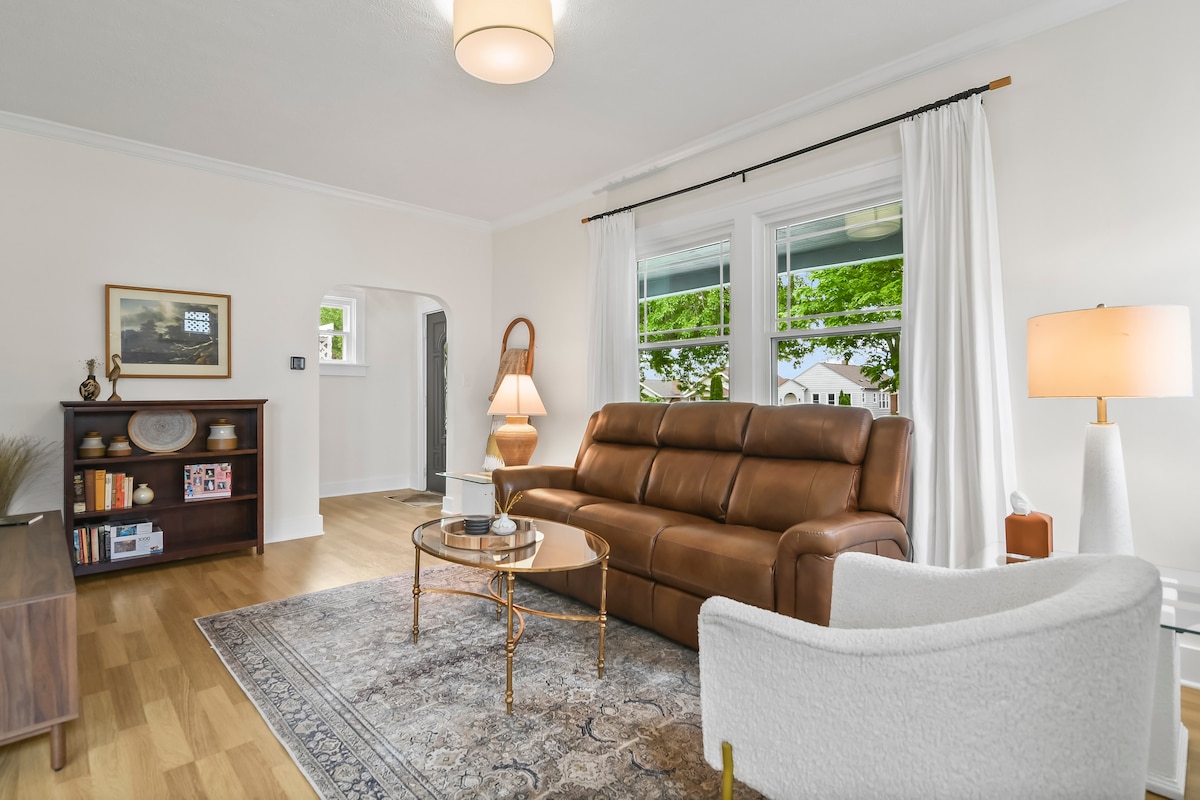 A spacious living area is featured, with a brown leather sofa facing a circular glass coffee table. Natural light fills the room through large windows, complemented by white curtains. A wooden shelf with decorative items and a floor lamp add warmth to the neutral-toned decor.