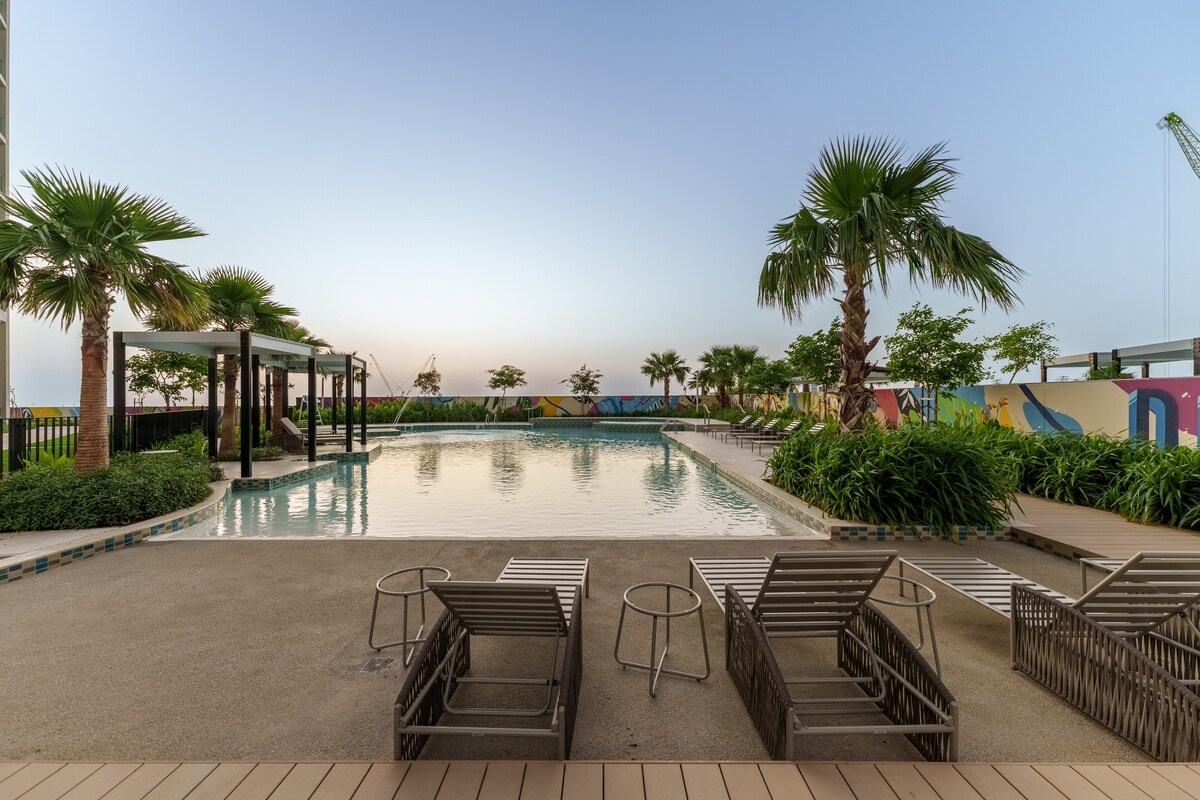 An outdoor pool area is displayed, featuring landscaped greenery and palm trees. Several lounge chairs are arranged around the poolside. The water glimmers under the soft evening light, creating a serene atmosphere. A shaded seating area is visible in the background, enhancing relaxation options.