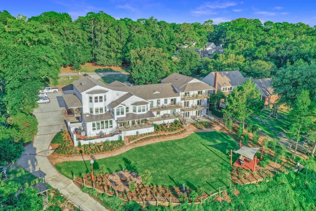 Aerial view of the Jubilee Suites complex showcases a spacious green lawn surrounded by gardens, with several parking spaces visible. The building features multiple balconies and a roofline that complements the natural landscape of trees in the background.