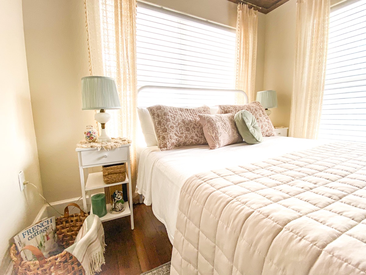 The cozy sleeping area features a spacious bed adorned with soft bedding and decorative pillows. Natural light filters through sheer curtains, illuminating the warm wooden floors. A bedside table holds a lamp, while a woven basket and a book are positioned nearby.