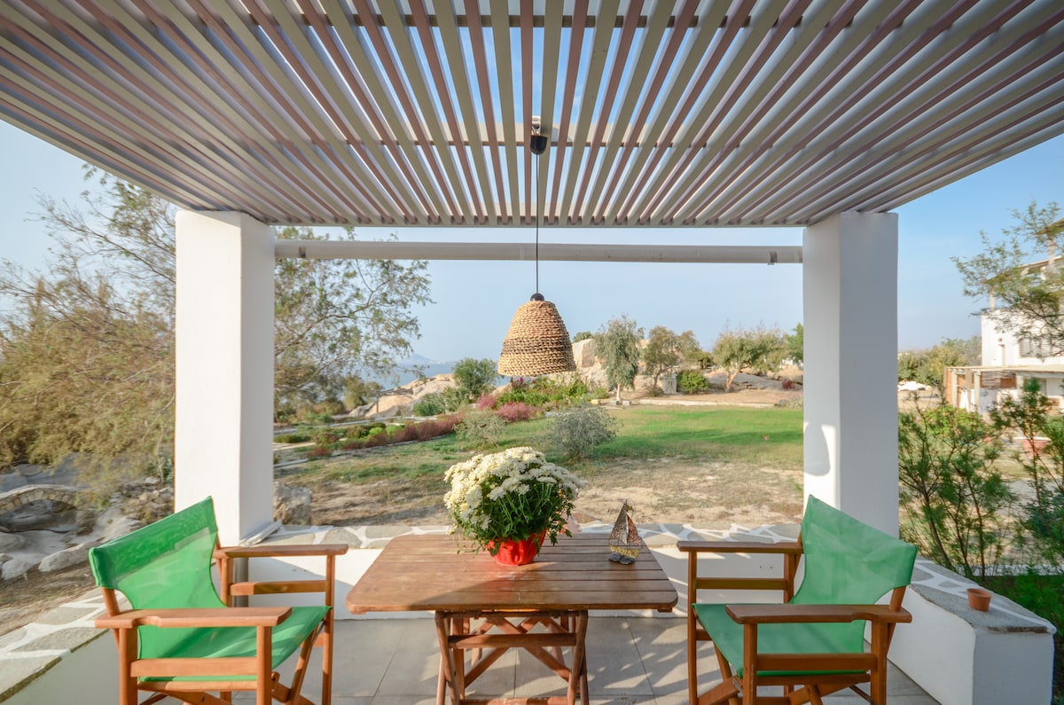 A shaded outdoor seating area is featured under a slatted pergola. Two green chairs with wooden accents surround a wooden table, complemented by a wicker pendant light overhead. A planter with white flowers sits on the table, with a scenic view of greenery and the sea visible in the background.