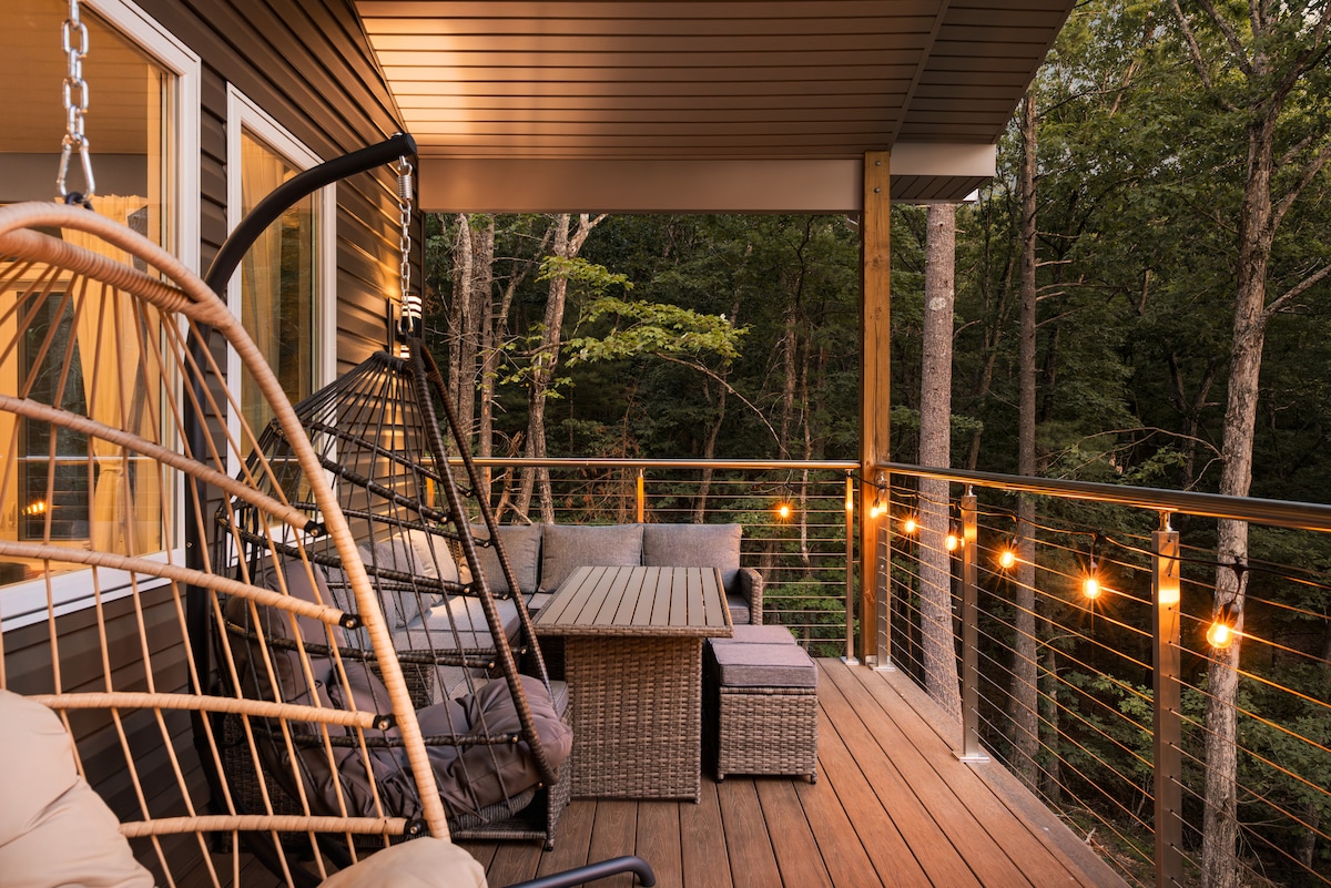 A spacious outdoor deck features comfortable seating areas, including a cozy rattan sofa and a hanging chair. Soft lights illuminate the space, creating a relaxed ambiance. The surrounding trees provide a sense of privacy and connect the area with nature.