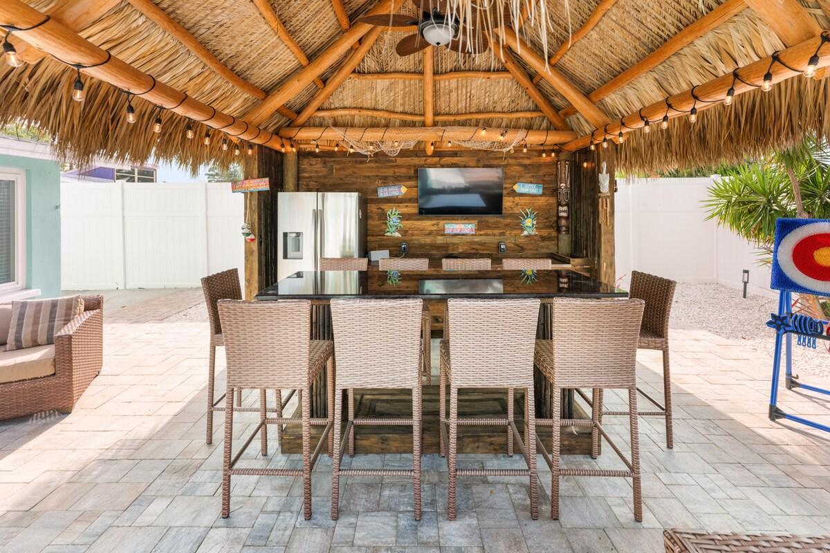 A Tiki-style bar area is showcased, featuring a thatched roof and a long counter with several high-backed chairs. Decorative lights illuminate the space, while a flat-screen TV is mounted on the wooden wall behind the bar. An inviting atmosphere for relaxation and socializing is created.