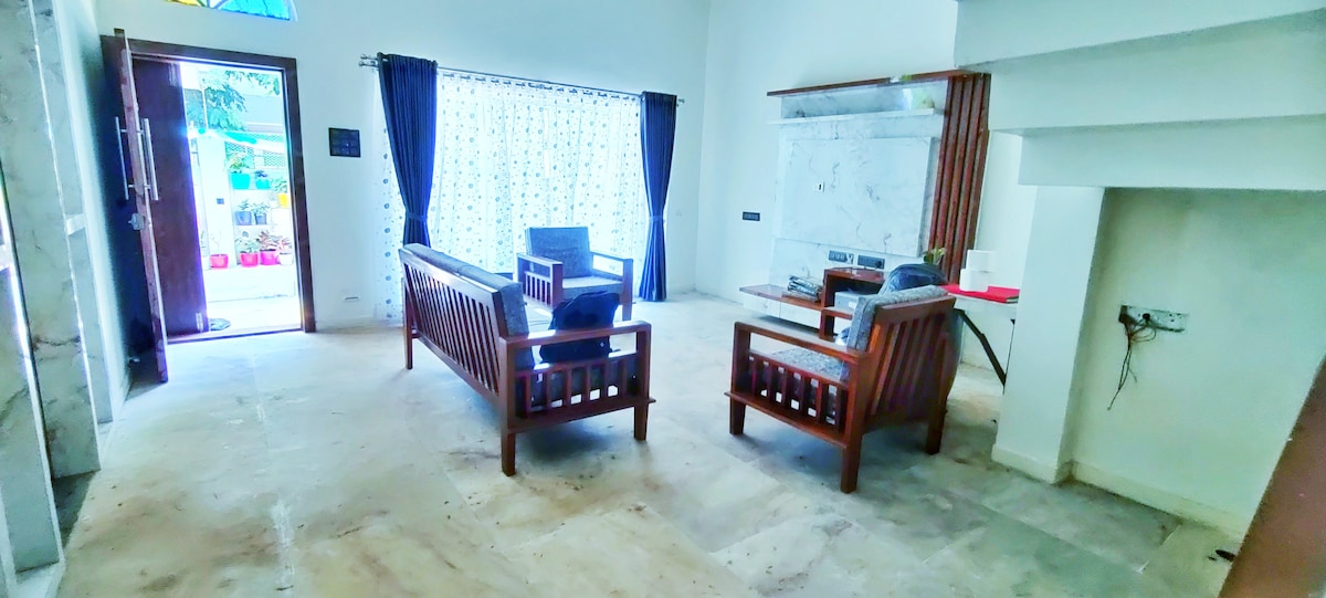 A well-lit living area features modern wooden furnishings, including a pair of armchairs and a couch. A coffee table is positioned centrally, with a decorative piece on top. Natural light filters through the sheer curtains, enhancing the bright ambiance of the space.