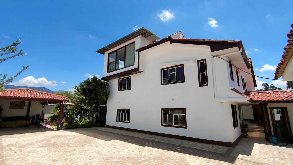 The exterior of the house is showcased, featuring a two-story structure with a modern design. Large windows allow natural light to fill the interior. The surrounding outdoor space includes a paved area and nearby greenery under a clear blue sky.