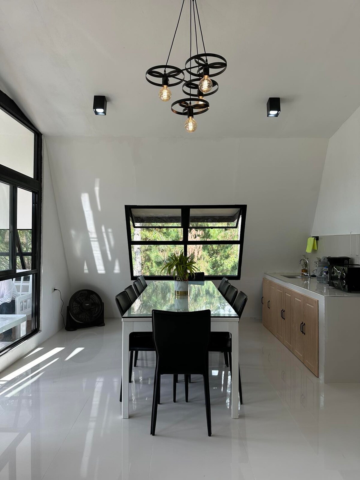 A modern dining area features a sleek glass table surrounded by six black chairs. A distinctive overhead light fixture is showcased, and large windows allow natural light to fill the space, illuminating the white walls and polished flooring.