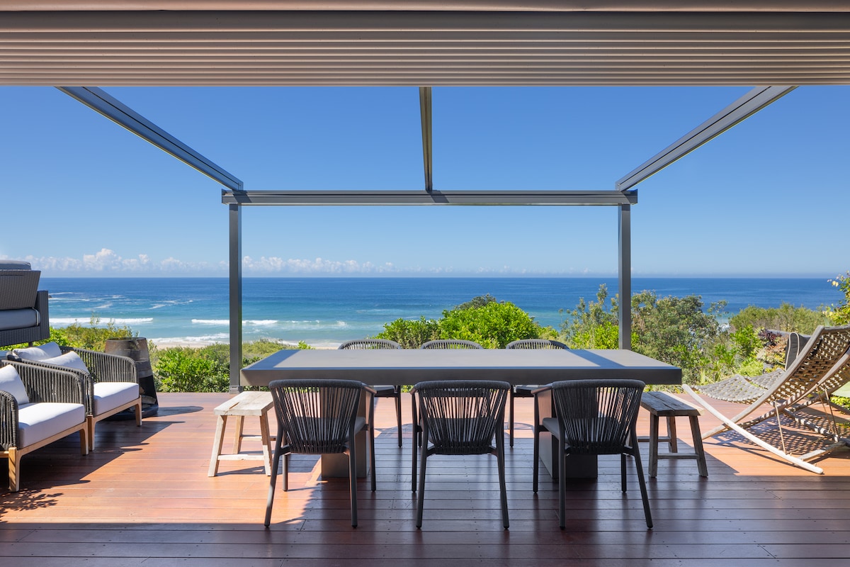An outdoor entertaining area features a spacious table surrounded by black chairs, with ocean views visible beyond a wooden deck. The space is partially covered by a pergola, enhancing the connection to the surrounding landscape and inviting natural light.