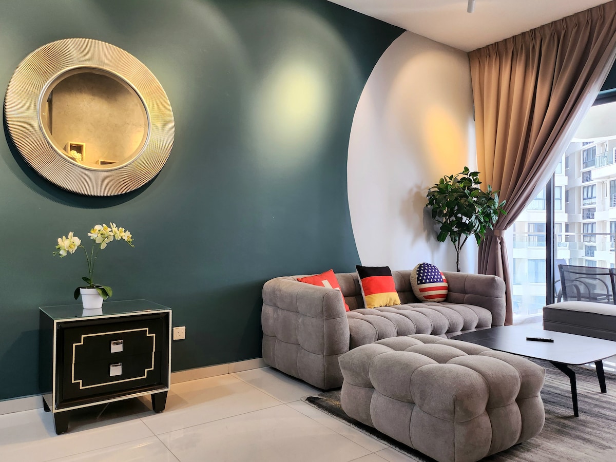 A modern living space is presented with a curved green accent wall and a full-length window allowing natural light. A plush sectional sofa in light gray features colorful cushions. A circular mirror and a small side table with drawer add to the stylish decor. Potted plant enhances the ambiance.