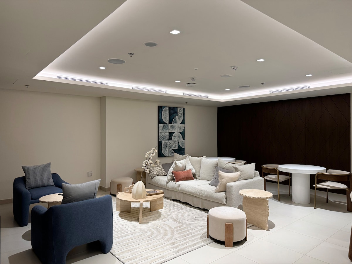 A spacious living area is depicted, featuring a light-colored sectional sofa with various cushions. Two blue accent chairs are positioned nearby, alongside natural wood side tables. A round dining table with wooden chairs is visible, complemented by stylish decor on the walls and a soft area rug.