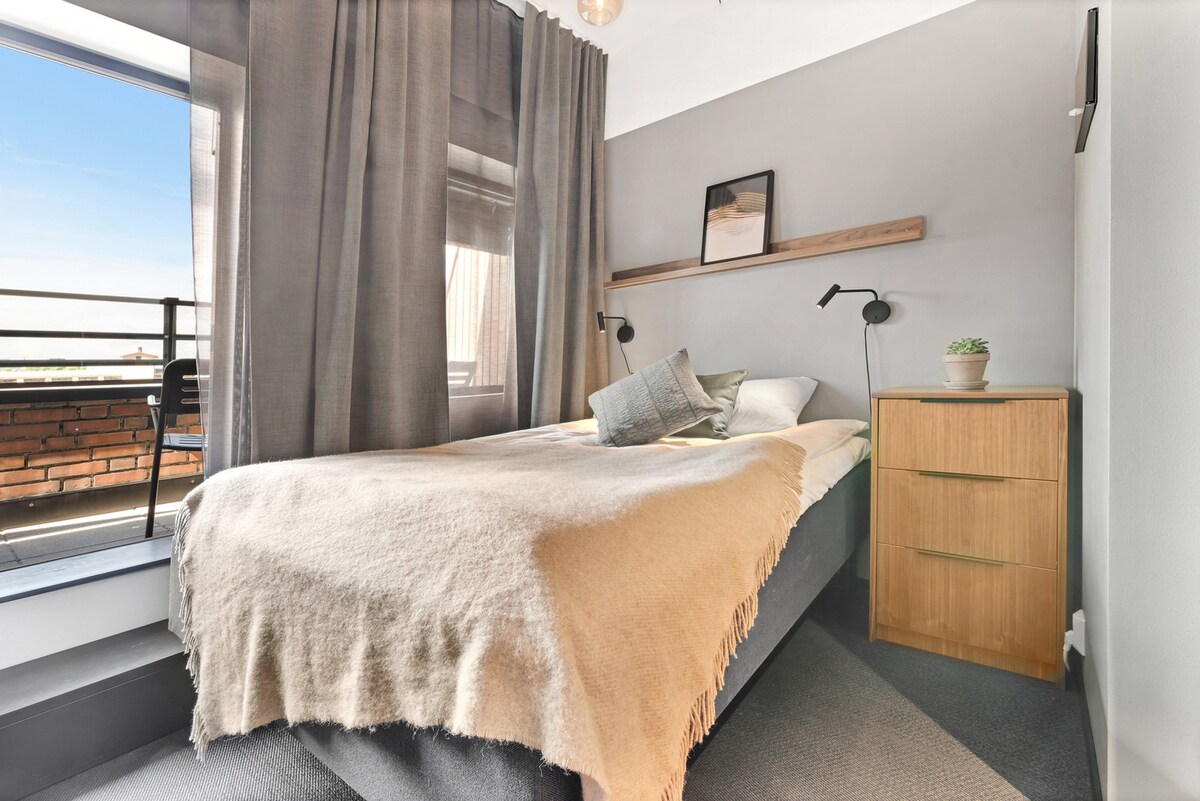 A small studio sleeping area features a spacious bed draped with a light, textured blanket. Soft lighting from two bedside lamps illuminates the room. Large windows provide access to fresh air and natural light, with sheer curtains partially filtering the view.