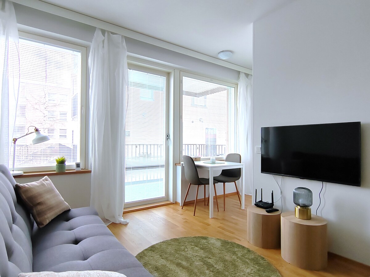 A bright living area features large windows with sheer curtains, allowing natural light to fill the space. A comfortable sofa is positioned beside a modern TV mounted on the wall. A dining table for two is set near the windows, accompanied by stylish chairs.