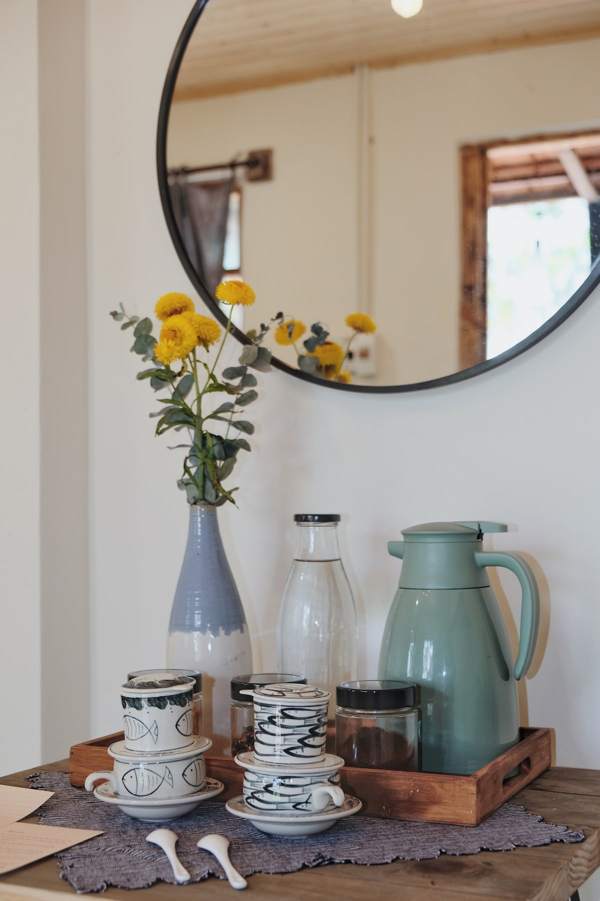 A round mirror reflects a collection of drinkware displayed on a wooden tray. Included are patterned mugs and a teapot alongside a glass jar, a pitcher, and a vase holding yellow flowers. A soft fabric lays beneath the arrangement, adding texture.