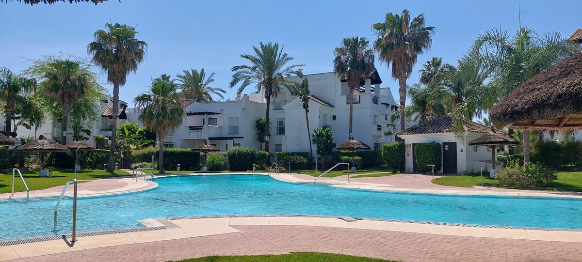 A communal swimming pool is featured, surrounded by well-maintained tropical gardens. Palm trees provide shade around the area, and thatched-roof cabanas are positioned alongside the pool. The clear blue water reflects the bright sky, creating a serene environment for relaxation.