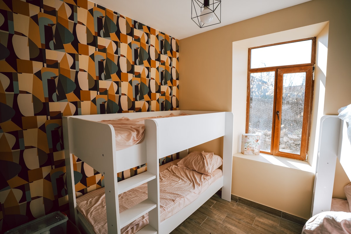 A well-organized dormitory features stylish bunk beds with soft bedding, set against a modern patterned accent wall. Natural light enters through a large window with wooden frames, illuminating the room's neutral tones and creating a comfortable environment.