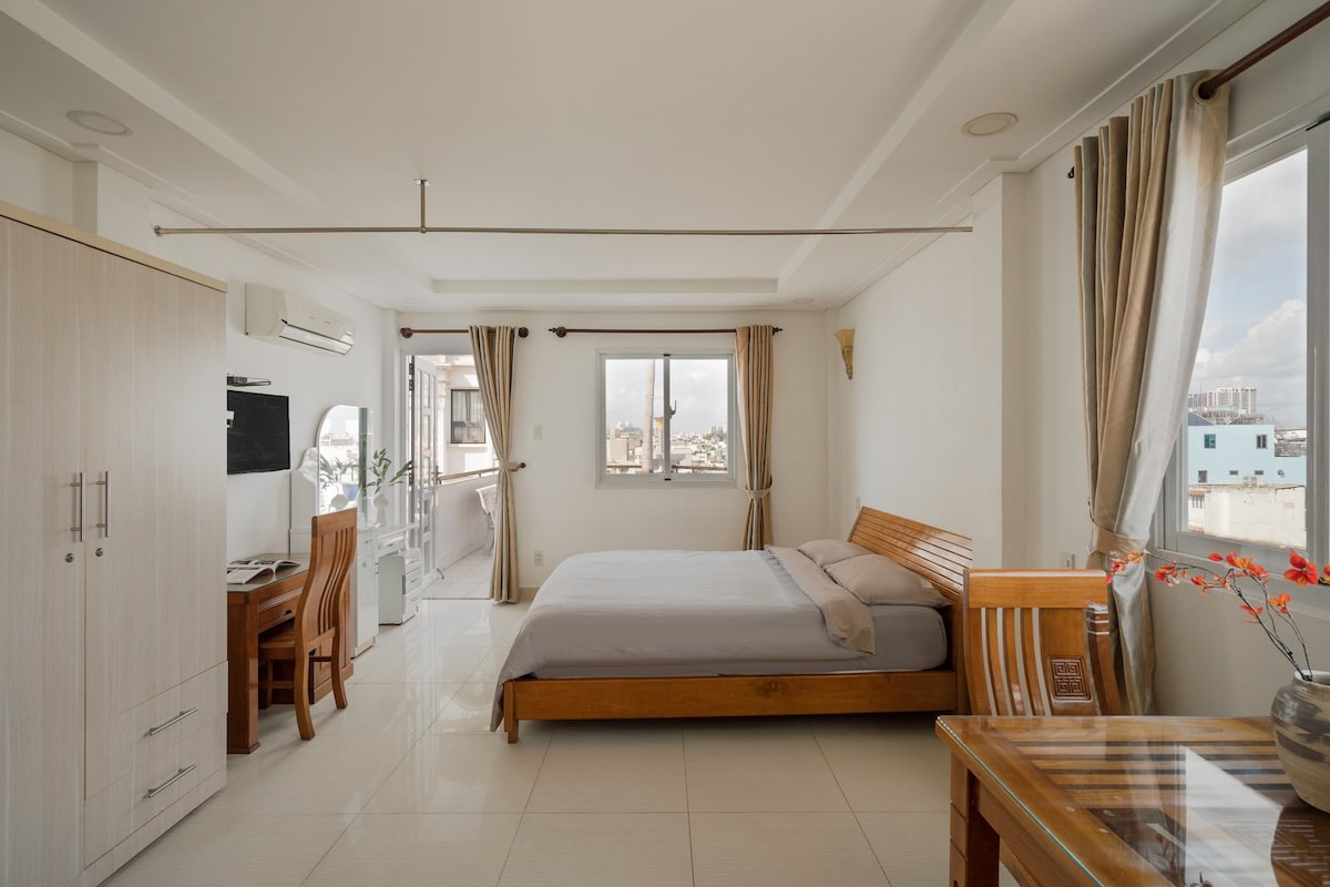A bright bedroom is complemented by large windows that allow natural light to fill the space. A wooden bed is centered in the room, accompanied by a simple desk and chair. Neutral walls are paired with elegant curtains, creating a tranquil atmosphere.