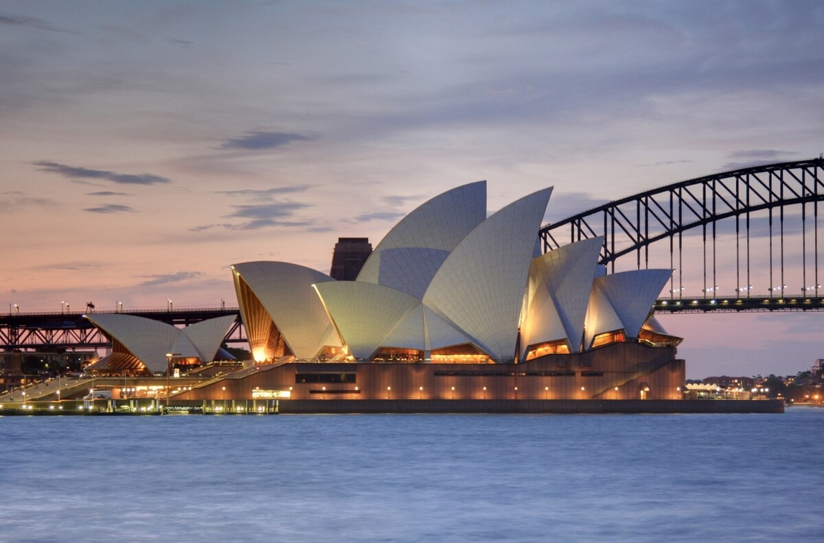 The Sydney Opera House is prominently featured, showcasing its distinctive sail-like design illuminated against the evening sky. Nearby, the Sydney Harbour Bridge stands in the background, completing the iconic skyline. The calm waters reflect the lights of the structure, enhancing the tranquil scene.
