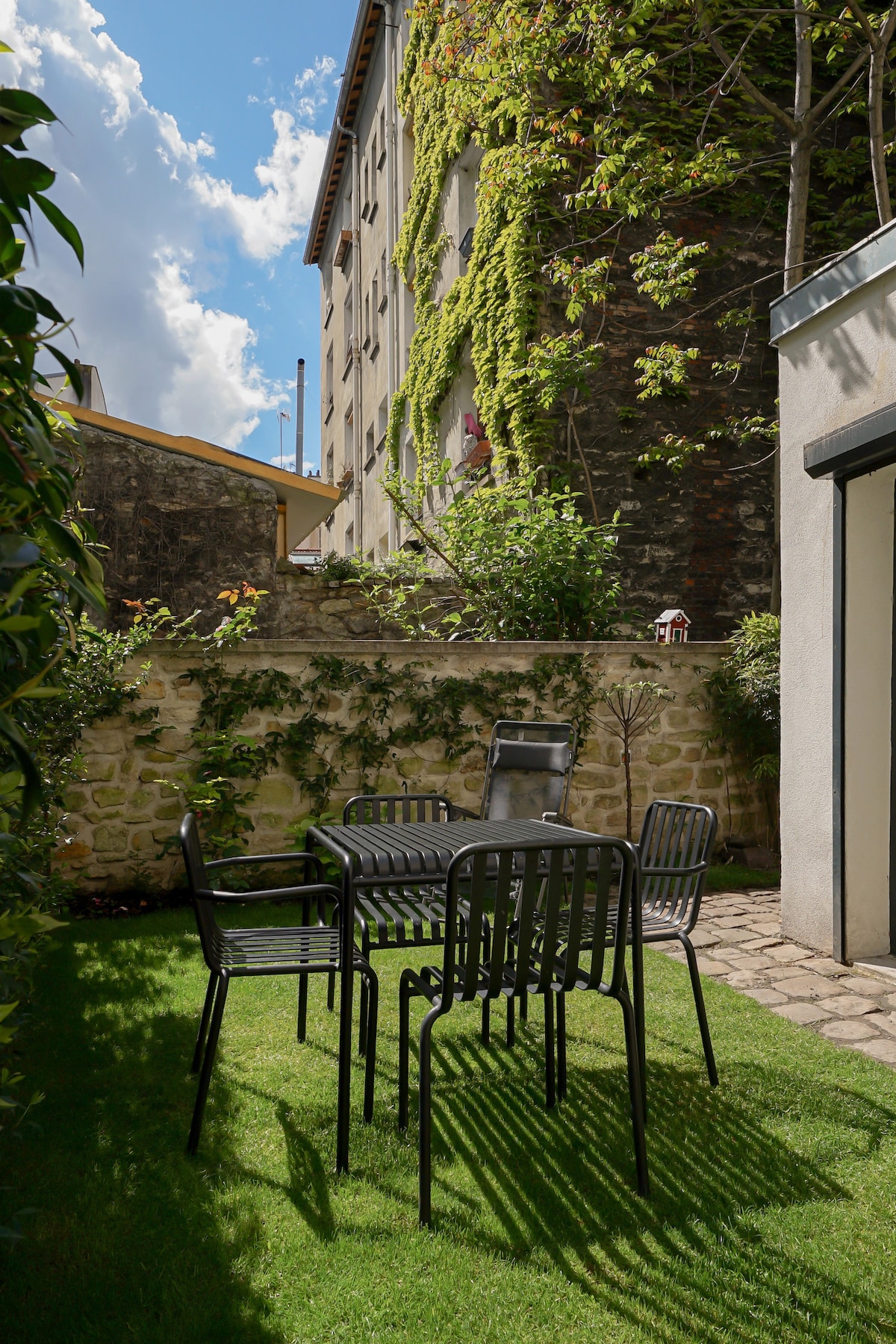 A small terrace is presented with a dark metal table surrounded by four chairs, set on a grassy area. A stone wall covered in green vines forms the background, while sunlight casts shadows over the space, contributing to a peaceful outdoor atmosphere.
