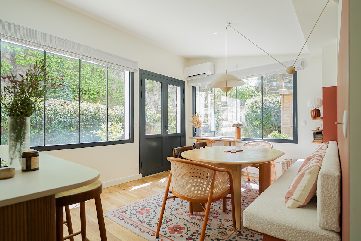 A bright dining area features large windows that overlook a green garden. A circular wooden table is surrounded by four chairs, and a cozy bench with a patterned cushion lines one side. Natural light illuminates the space, highlighting the warm wood floors.