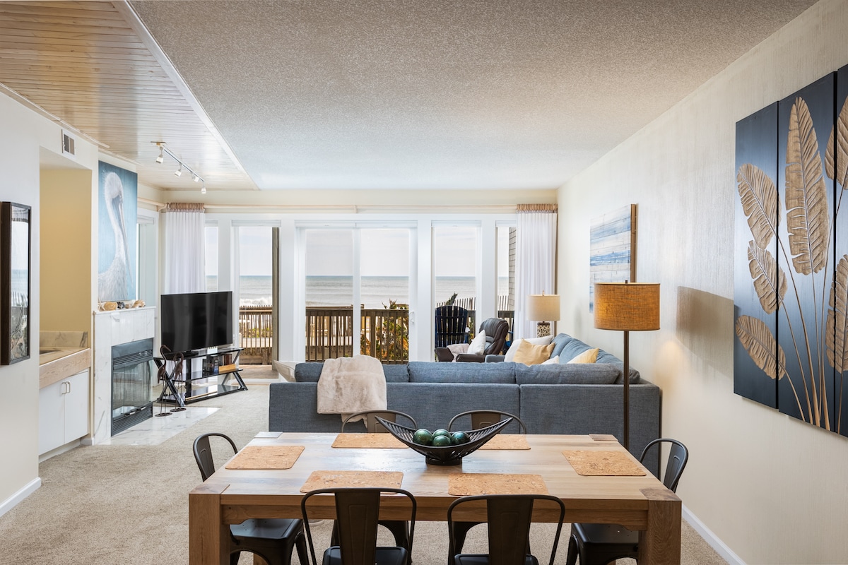 An open living area is showcased, featuring a spacious couch overlooking large windows with ocean views. A dining table with six chairs is positioned nearby, and a flat-screen television is visible in the corner. Soft natural light fills the room, creating a bright ambiance.