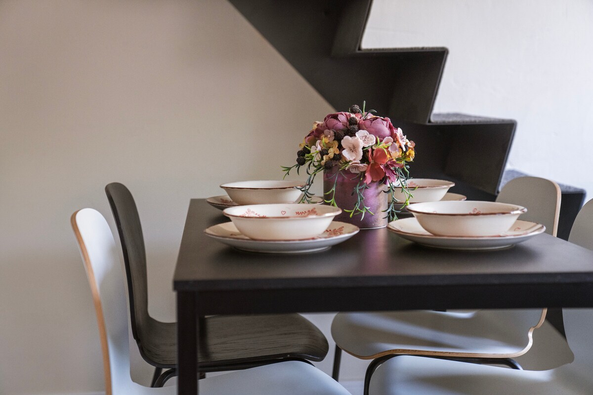 A dining table is set with five bowls arranged symmetrically, accompanied by a decorative vase filled with colorful flowers. The table is surrounded by stylish black and white chairs, with a staircase visible in the background.