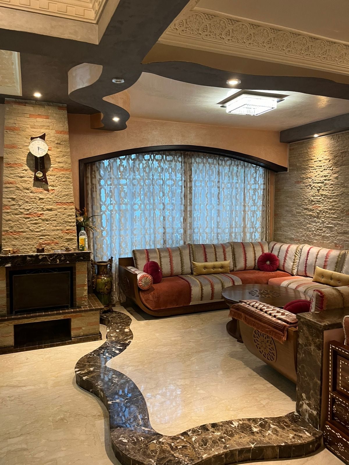 A spacious living area features a curved sofa adorned with colorful cushions, while natural light filters through patterned curtains. A decorative stone fireplace is positioned against a textured wall, complemented by marble flooring that adds an elegant touch.