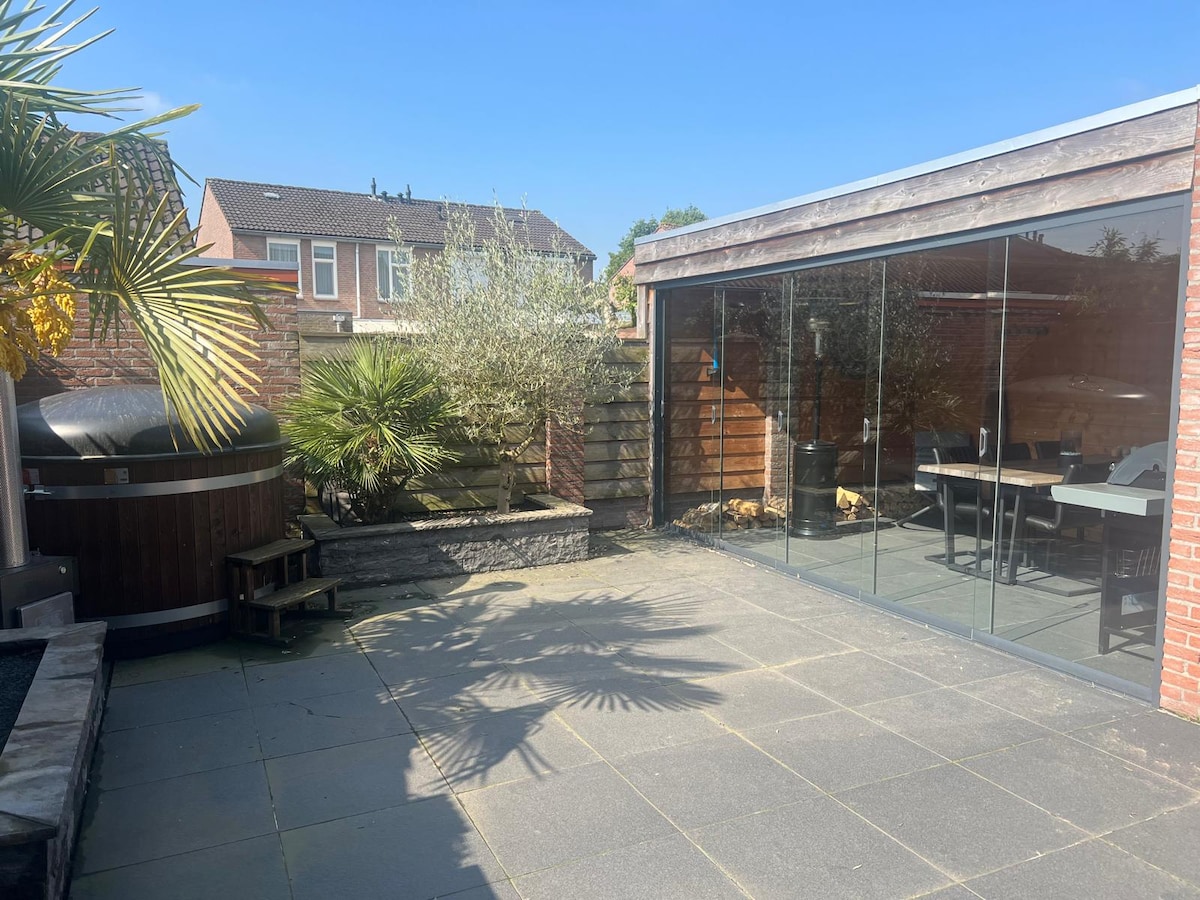 A spacious outdoor area features modern stone paving and a neatly landscaped garden. Glass doors provide access to the interior, while a wooden structure houses a hot tub, surrounded by greenery and a table with chairs. Sunshine brightens the serene atmosphere.