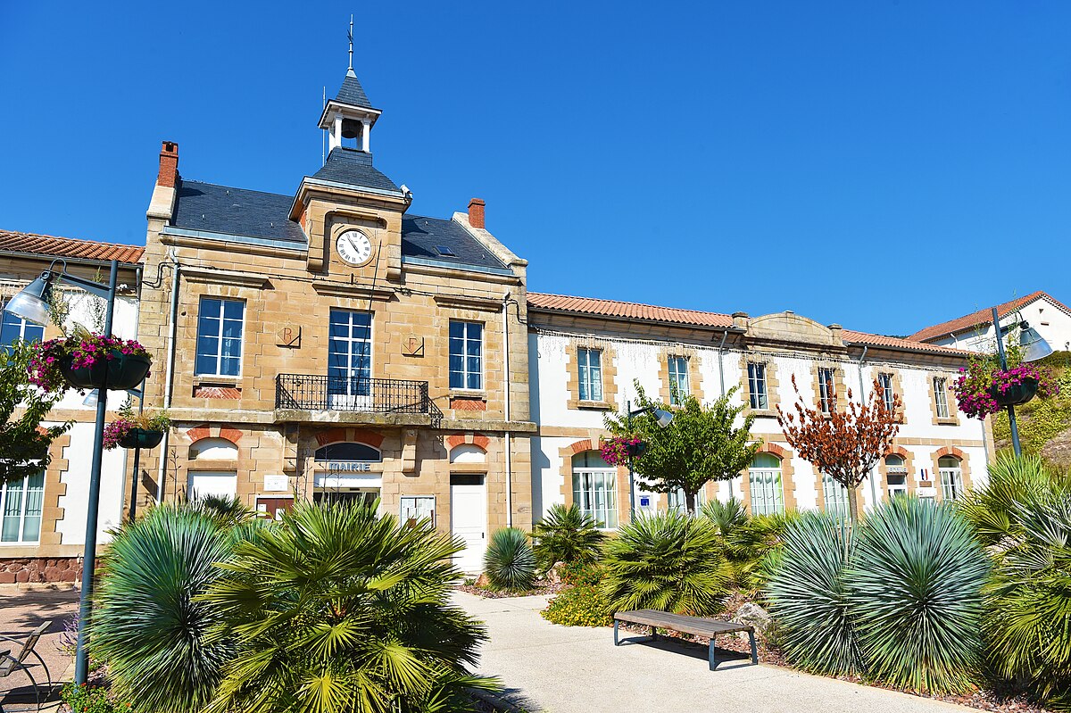 A historical building features a central clock tower topped with a spire. Surrounding greenery includes palm plants and well-maintained landscaping. The structure displays a mixture of traditional and modern architectural elements, with large windows and a welcoming entrance flanked by decorative plants.