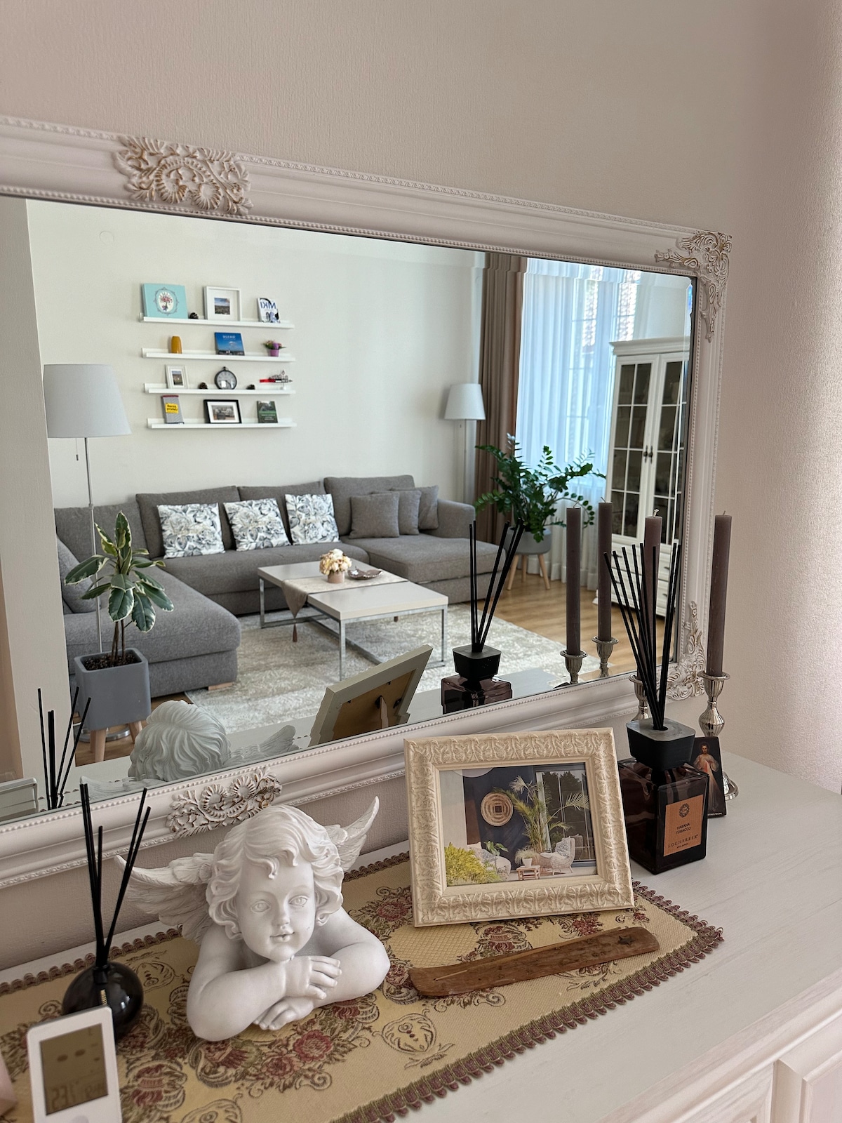 A stylish living room is reflected in a large ornate mirror. A cozy gray sofa is paired with patterned cushions. A coffee table sits in the center, surrounded by greenery from potted plants, while decorative items, including candles and a photo frame, are arranged on a side table.