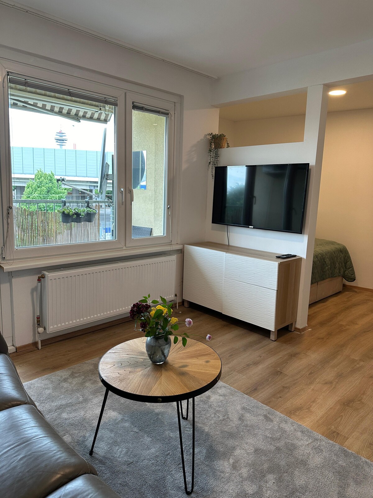 A living area is presented with a comfortable sofa and a small round table topped with a flowering plant. A television is mounted on the wall opposite, and large windows provide views of greenery outside.