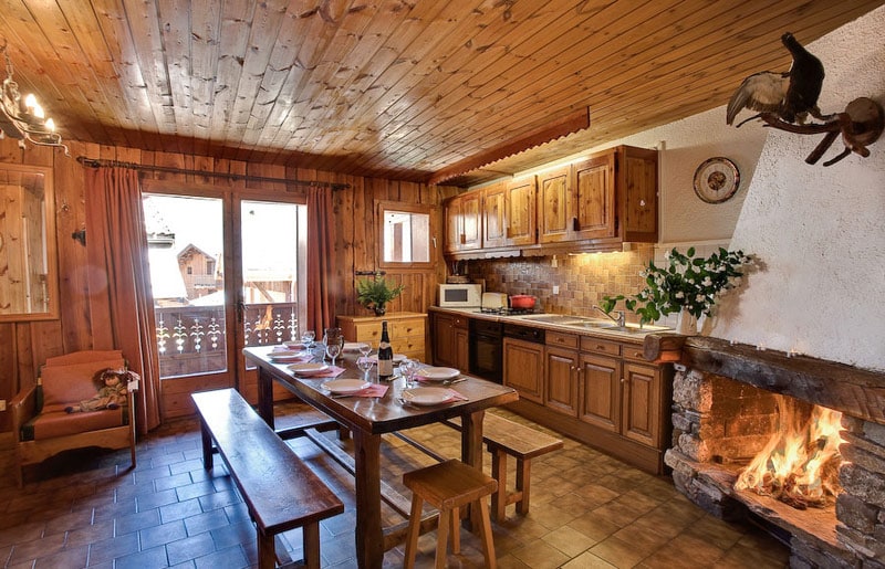 The dining area features a wooden table set for guests, complemented by benches and a cozy armchair. A fireplace adds warmth to the space, while wooden cabinetry and stone accents enhance the rustic ambiance. A large window opens to the balcony, inviting natural light.