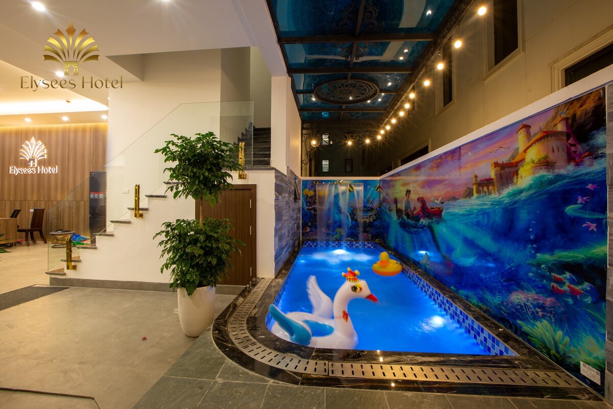 An indoor swimming pool is surrounded by vibrant murals depicting a beach scene. Decorative lighting is featured overhead, while a potted plant adds a touch of greenery. A staircase in the background leads to another level, and the hotel name is displayed on the wall.
