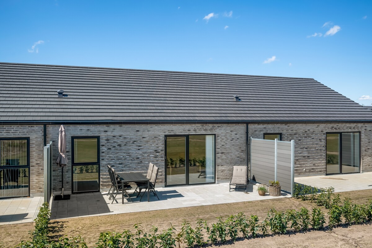 A modern row house is depicted, featuring a gray brick facade and large glass sliding doors. An outdoor patio area is shown with a table and chairs, along with a shaded umbrella. Green grass surrounds the area, highlighting the spacious surroundings.