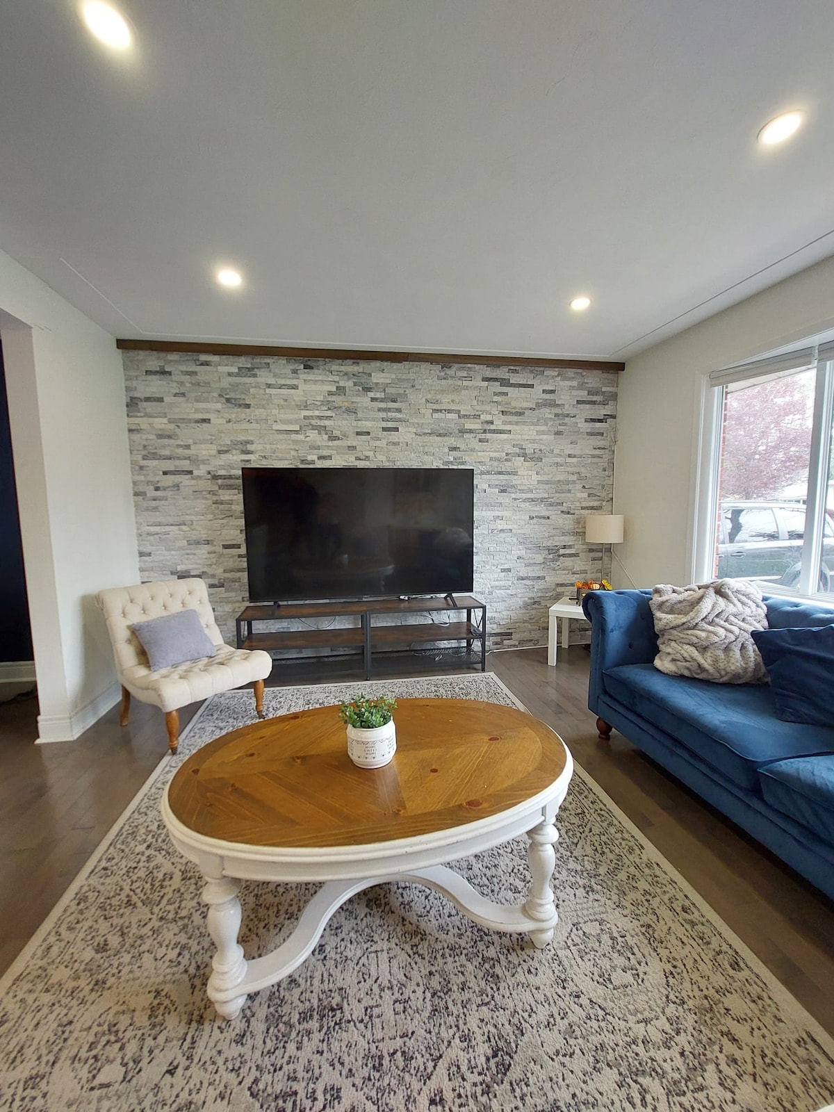 A living room features a large television mounted against a textured stone wall. A comfortable blue sofa sits adjacent to a round wooden coffee table. A small potted plant decorates the table, while natural light fills the space through large windows.
