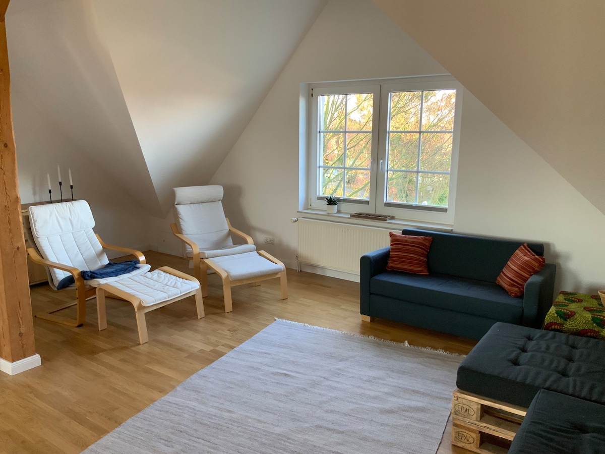 A light-filled living area features a dark sofa with decorative pillows and two white armchairs positioned beside a window. A neutral area rug is laid on the wooden floor, while natural light illuminates the room from the large window.