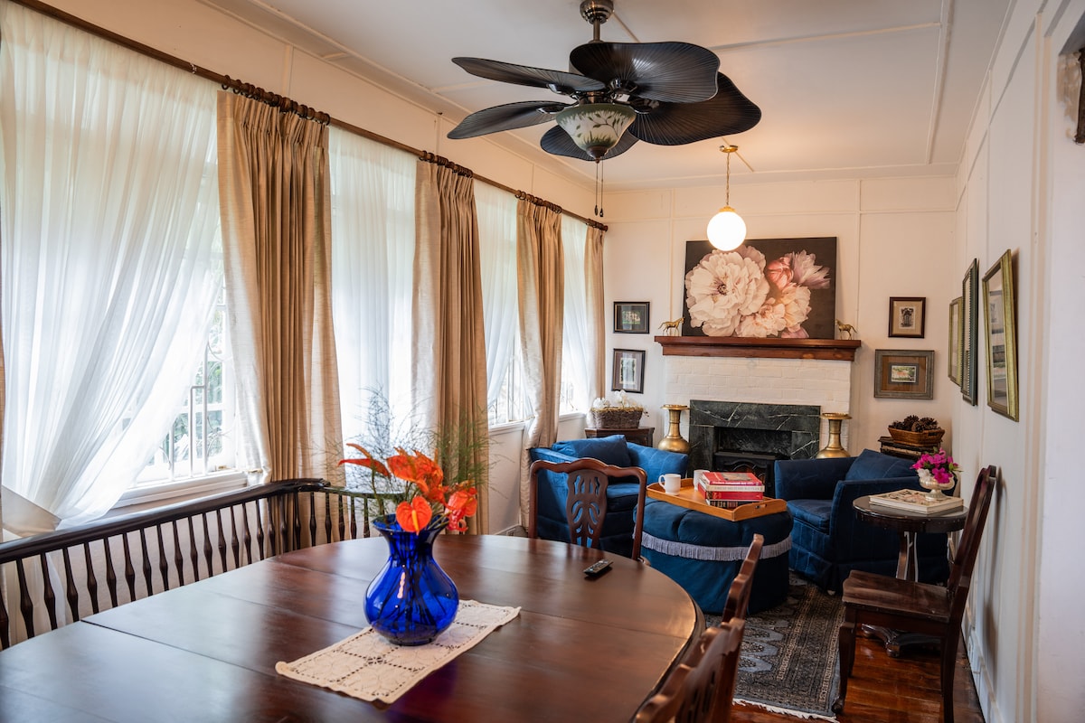 A cozy living area features a blue seating arrangement with a coffee table positioned in front. Natural light fills the space through large, flowing curtains. A decorative vase with flowers sits on a wooden dining table, while a wall art piece adds character to the room.