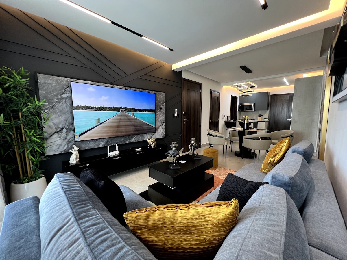 A spacious living area is presented, featuring a modern, light gray sofa with decorative pillows. A large flat-screen TV displays a serene waterfront scene on a textured wall. A coffee table anchors the space, while a dining area is visible in the background.