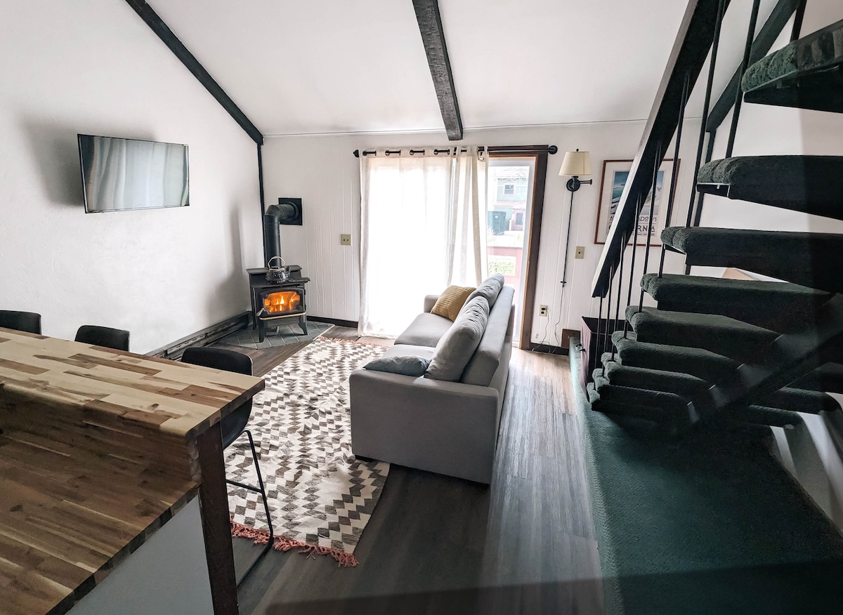 A light-filled living area features a spacious sofa facing a modern television and a warm wood-burning stove. An area rug adds texture to the floor, while a staircase leads to the upper level. Large windows allow natural light to illuminate the room.