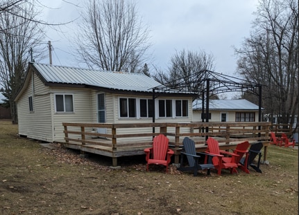 A cabin exterior is seen with a wraparound deck featuring several lounge chairs in red and black. Large windows provide natural light, and bare trees surround the area, indicating a tranquil setting. A nearby structure is partially visible in the background.