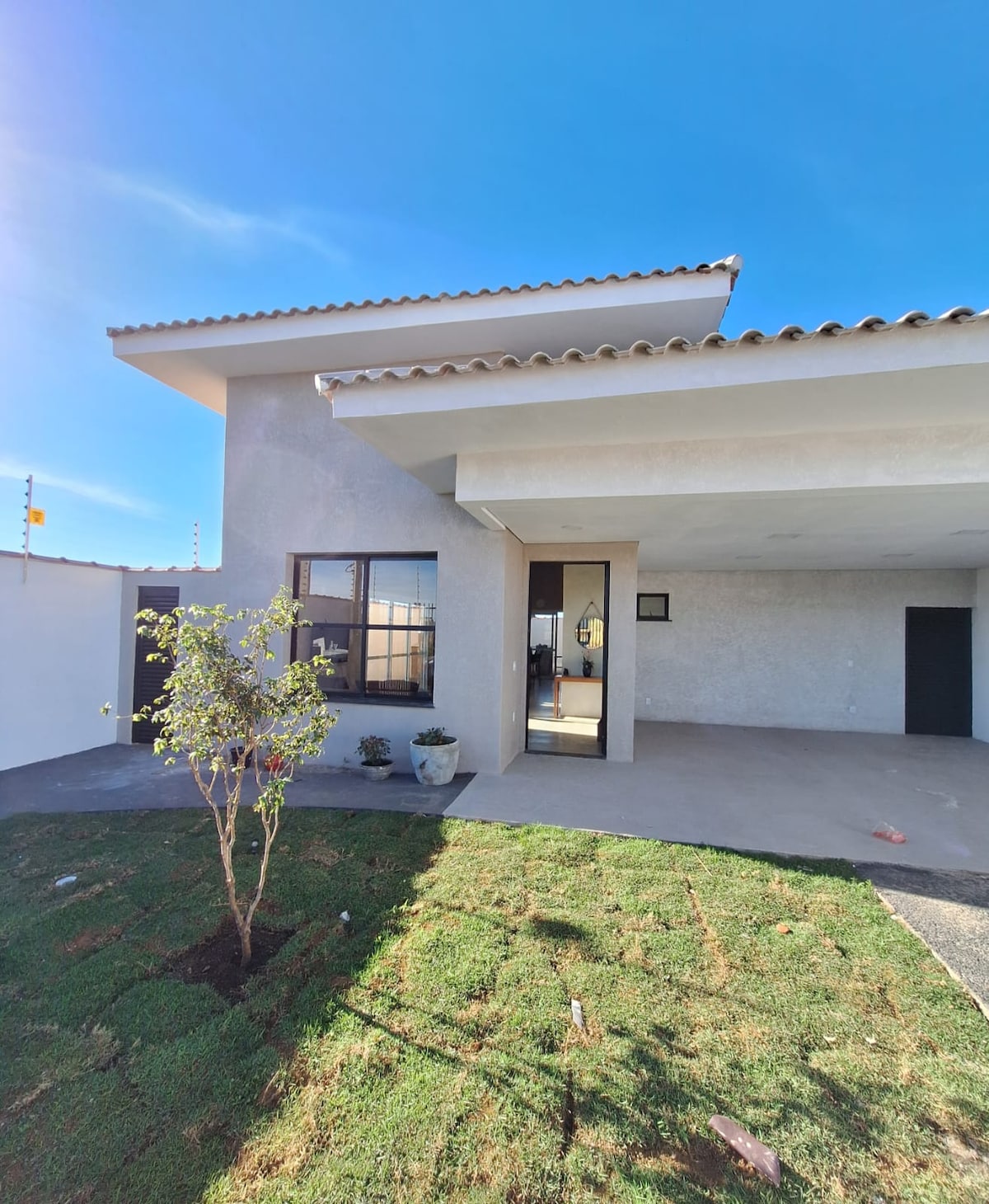 A modern house exterior features a clean, minimalist design, with angled rooflines and a light-colored façade. A small tree and potted plant are situated in a well-maintained yard, while a spacious entrance leads into the interior of the home.