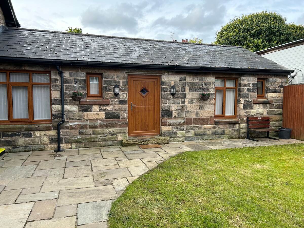 The exterior of The Coachman's Cottage features a charming stone façade with wooden accents. A sturdy front door is flanked by flower pots, while neatly laid stone pathways lead to a spacious green lawn, creating an inviting entrance to the property.