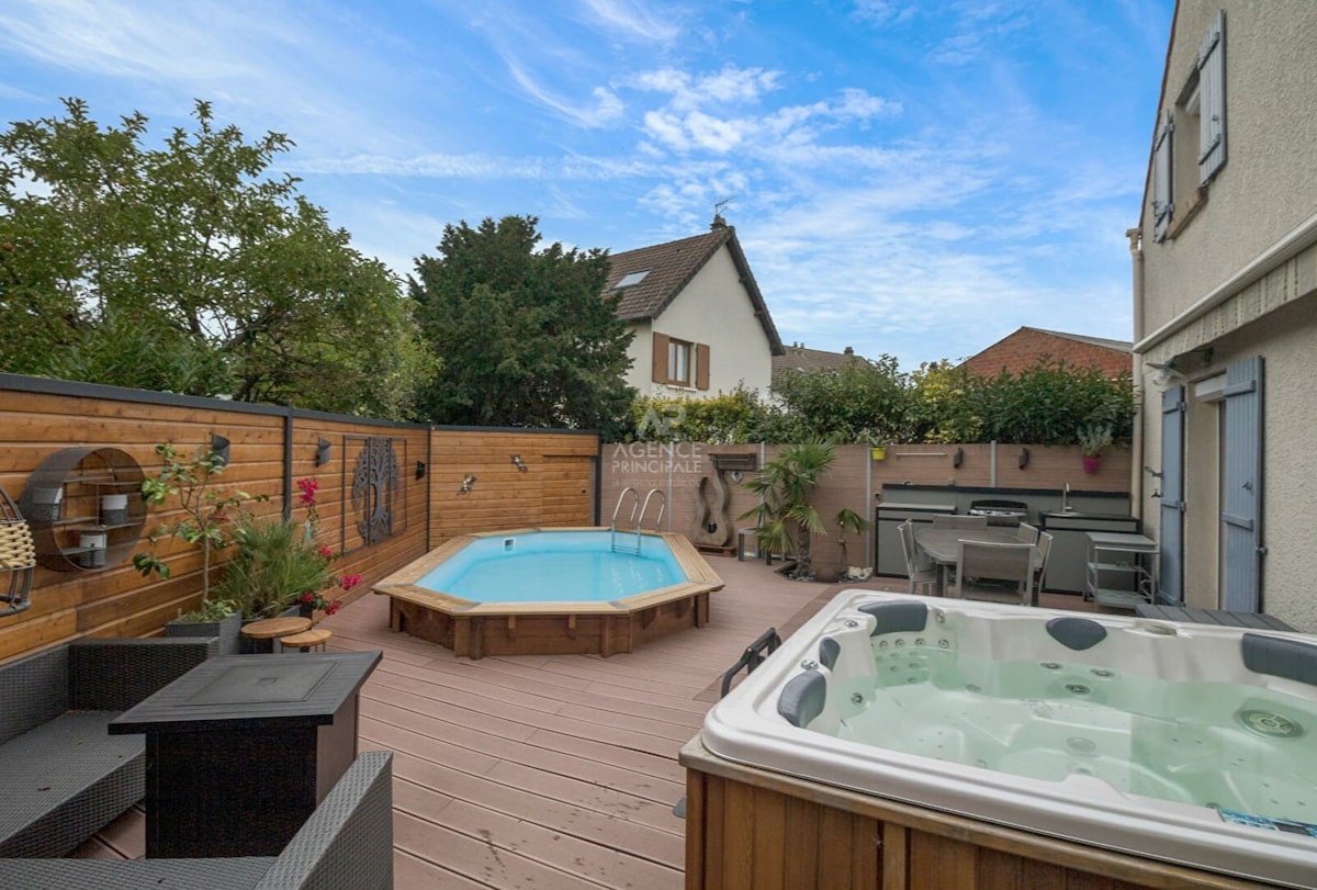 An outdoor space features a small wooden deck with a rectangular pool and a round hot tub positioned nearby. Comfortable seating arrangements are present next to lush greenery and decorative plants, creating an inviting environment for relaxation.