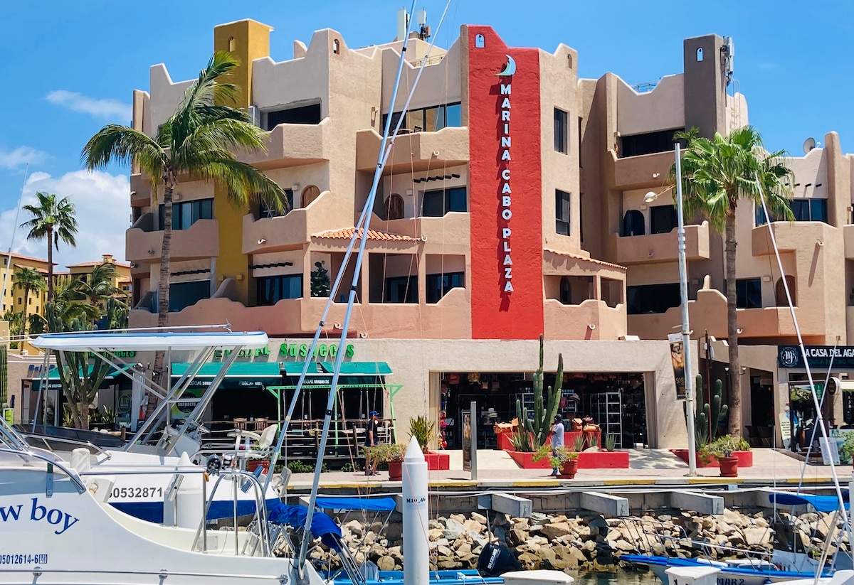The Marina Cabo Plaza building is prominently displayed along the waterfront, featuring a vibrant red sign. Palm trees frame the structure, while boats are docked in front, creating a lively marina scene. The surrounding area showcases a mix of shops and restaurants.