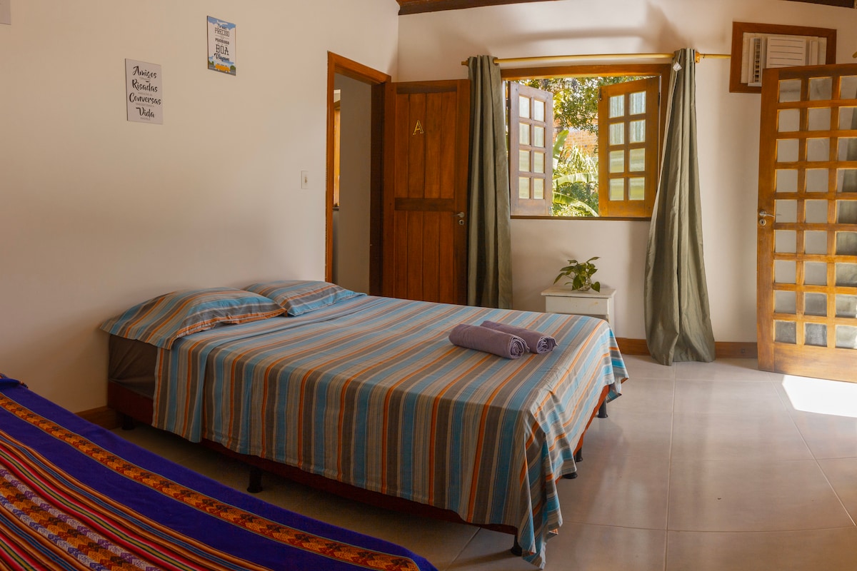 A bright sleeping area features a spacious bed with a striped blanket, accompanied by a smaller bed. Natural light filters through large windows dressed with green curtains. A potted plant sits on a side table, and wooden doors add warmth to the room's minimalist decor.