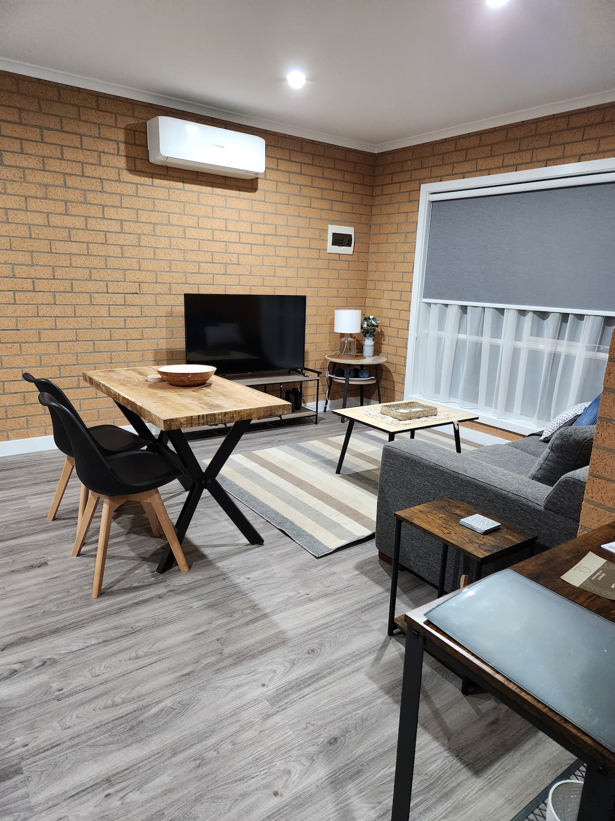 A cozy living area features a brick wall that complements the modern decor. A wooden dining table is paired with black chairs, while a small side table sits beside a comfortable gray sofa. A flat-screen TV is positioned across from the seating area.