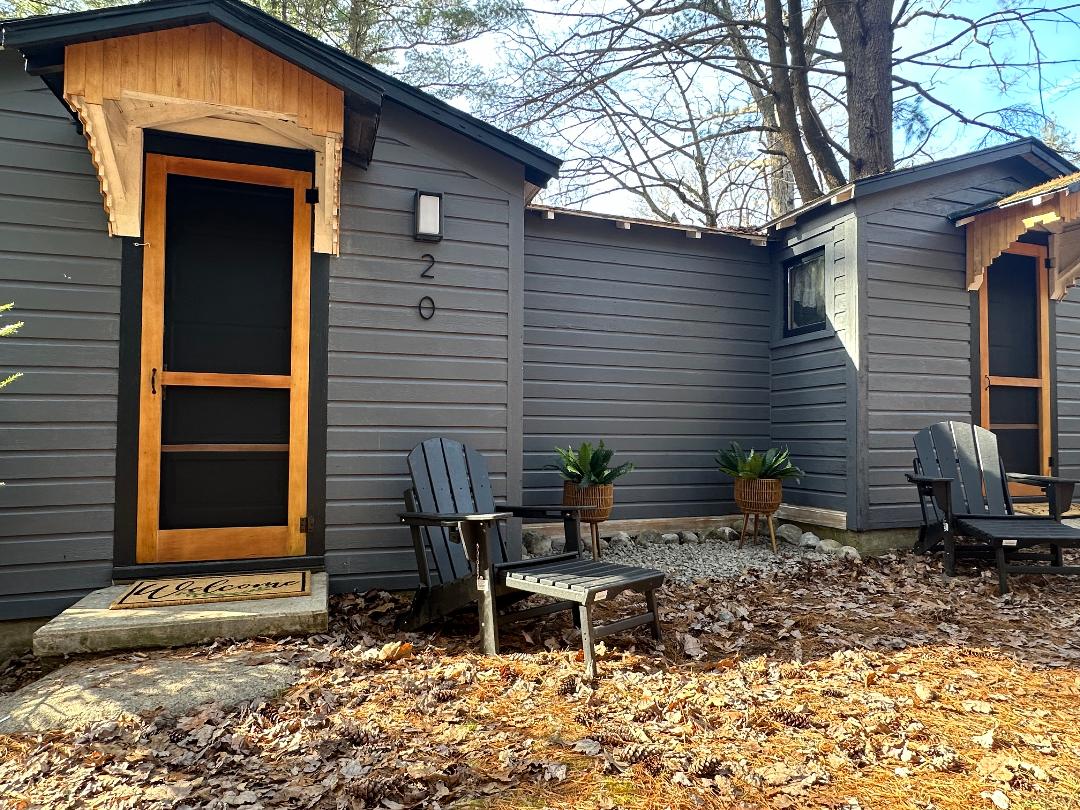 The exterior of a rustic cabin is shown, featuring a welcoming entrance with a wooden door. Two black Adirondack chairs are placed on a leaf-covered ground, flanked by small planters. The cabin is shaded by trees, providing a serene outdoor setting.