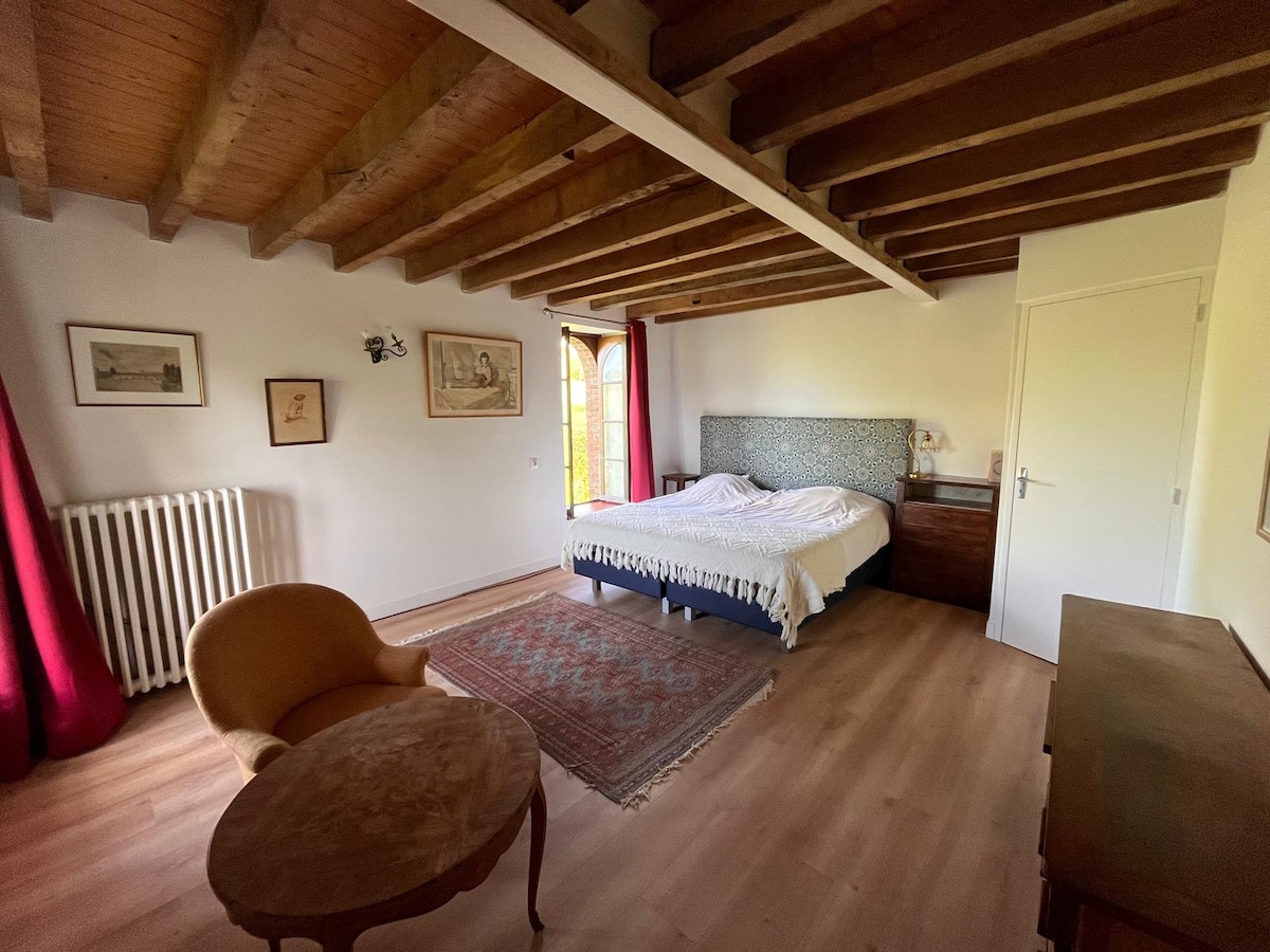A spacious bedroom features exposed wooden beams overhead and soft natural light coming from a large window with red curtains. A comfortable armchair is positioned beside a small round table, while the bed, adorned with a decorative headboard, adds an inviting touch to the overall layout.