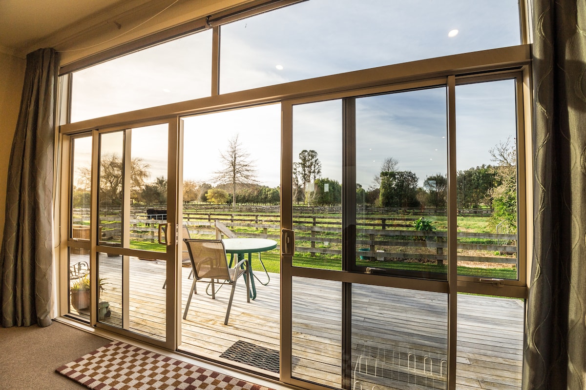Large glass doors open to a spacious deck that overlooks serene farmland. A table and chairs are arranged on the deck, inviting outdoor relaxation. The view features lush greenery and trees, enhancing the tranquil setting.